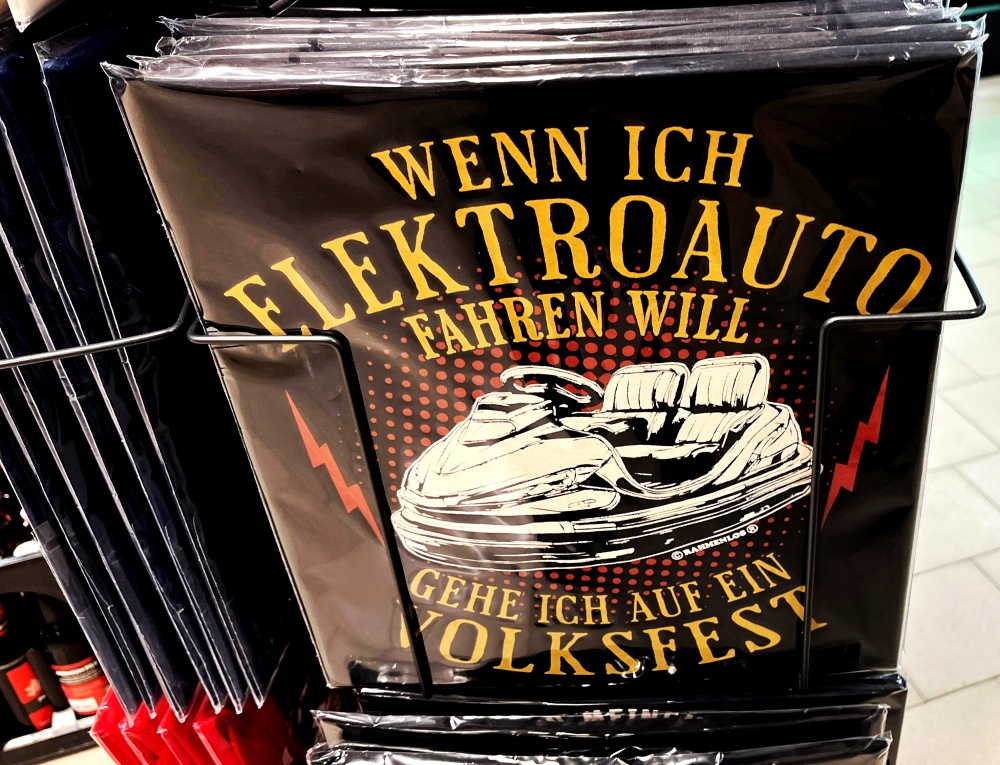 zeigt ein verpacktes T-Shirt im Supermarkt mit der Aufschrift 'Wenn ich Elektroauto fahren will, gehe ich auf ein Volksfest'